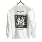A white crewneck sweatshirt hanging on a black hanger against a dark gray wall. The back of the sweatshirt features a large graphic print with a black and white patterned border, a large stylized 'M' logo in the center, and text reading 'ROY SROUF RT' above and 'ATLANTIC' below.