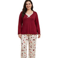 A woman modeling a two-piece sleepwear set in a bedroom. The set includes a long-sleeve burgundy top with a lace-up V-neck over white lace, and matching white pants with a red and pink floral print.