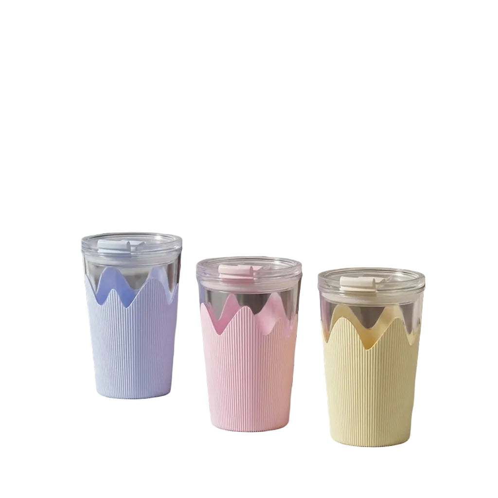 Three reusable cups with clear bodies and pastel ribbed sleeves in lavender, pink, and pale yellow, arranged on a tabletop in a modern indoor setting.