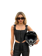 A woman stands on a dirt off-road track wearing a black lace-up corset-style top and black pants, holding a black and gray full-face helmet. Off-road vehicles are visible in the background under an overcast sky.