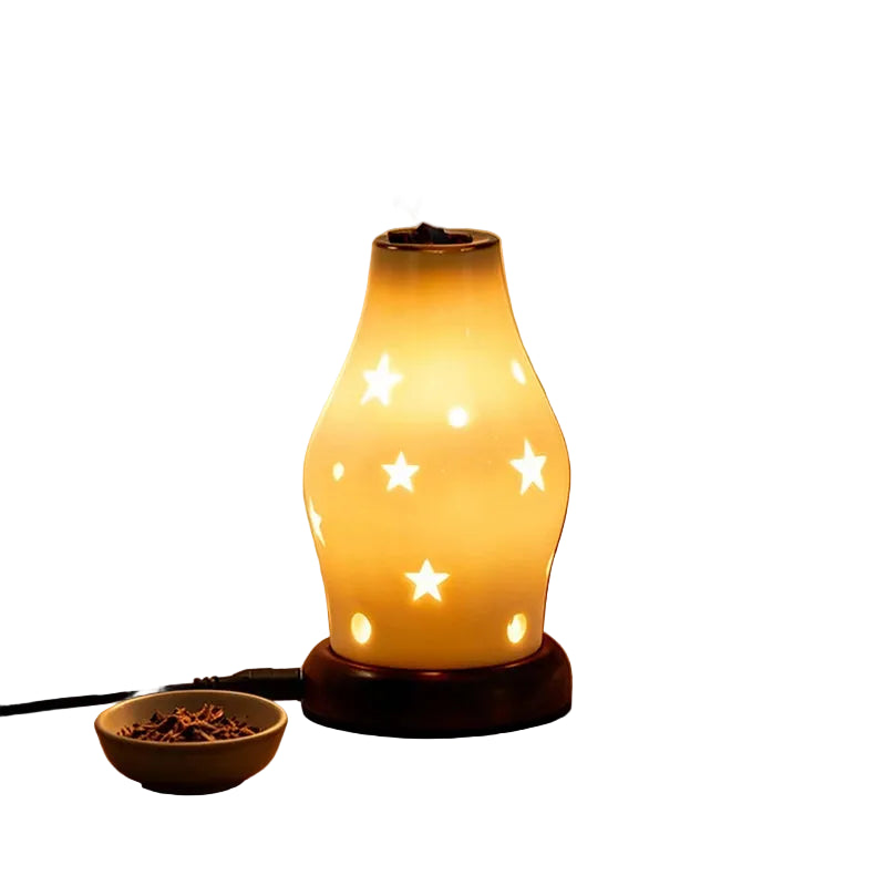 A glowing diffuser lamp with star-shaped cutouts sits on a wooden bedside table, casting star-shaped light projections on the wall. A small bowl of dried botanicals is placed beside it, with a bed and wooden headboard visible in the softly lit bedroom setting.