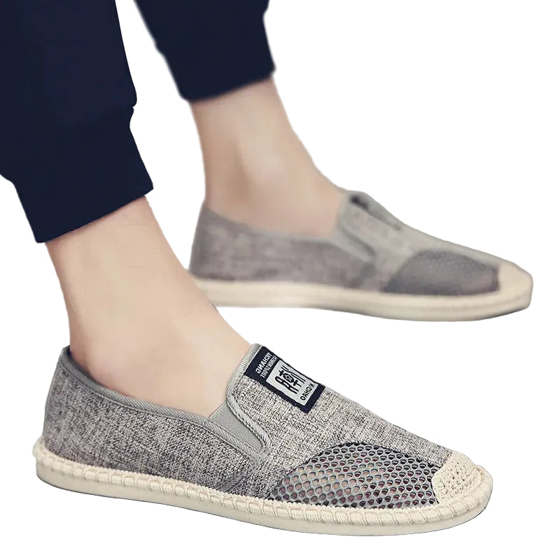 A person wearing gray slip-on mesh sneakers with a braided off-white sole, shown from the knees down against a light gray floor and wall.