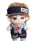 A small plush doll with light brown hair and large brown eyes, wearing a navy denim visor with a red apple patch, a blue-and-white checkered bib, and denim shorts with red plaid accents, posed against a plain cream wall.