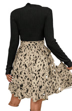 Back view of beige ribbed knit A-line dress with black ribbed long sleeves and patterned skirt with waist tie