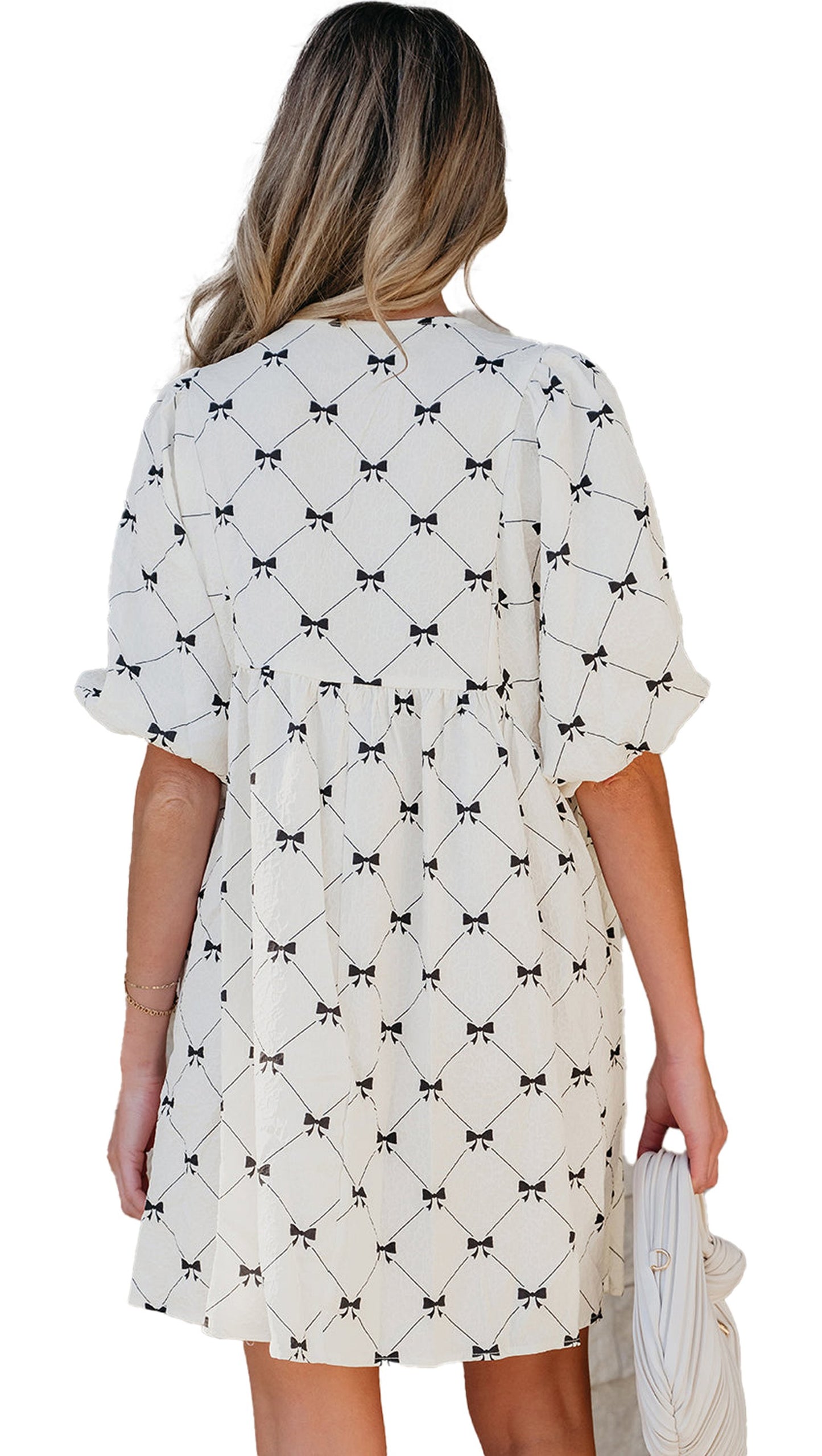Back view of model wearing white bowtie mini dress with black bow pattern short sleeves gathered waist holding white clutch