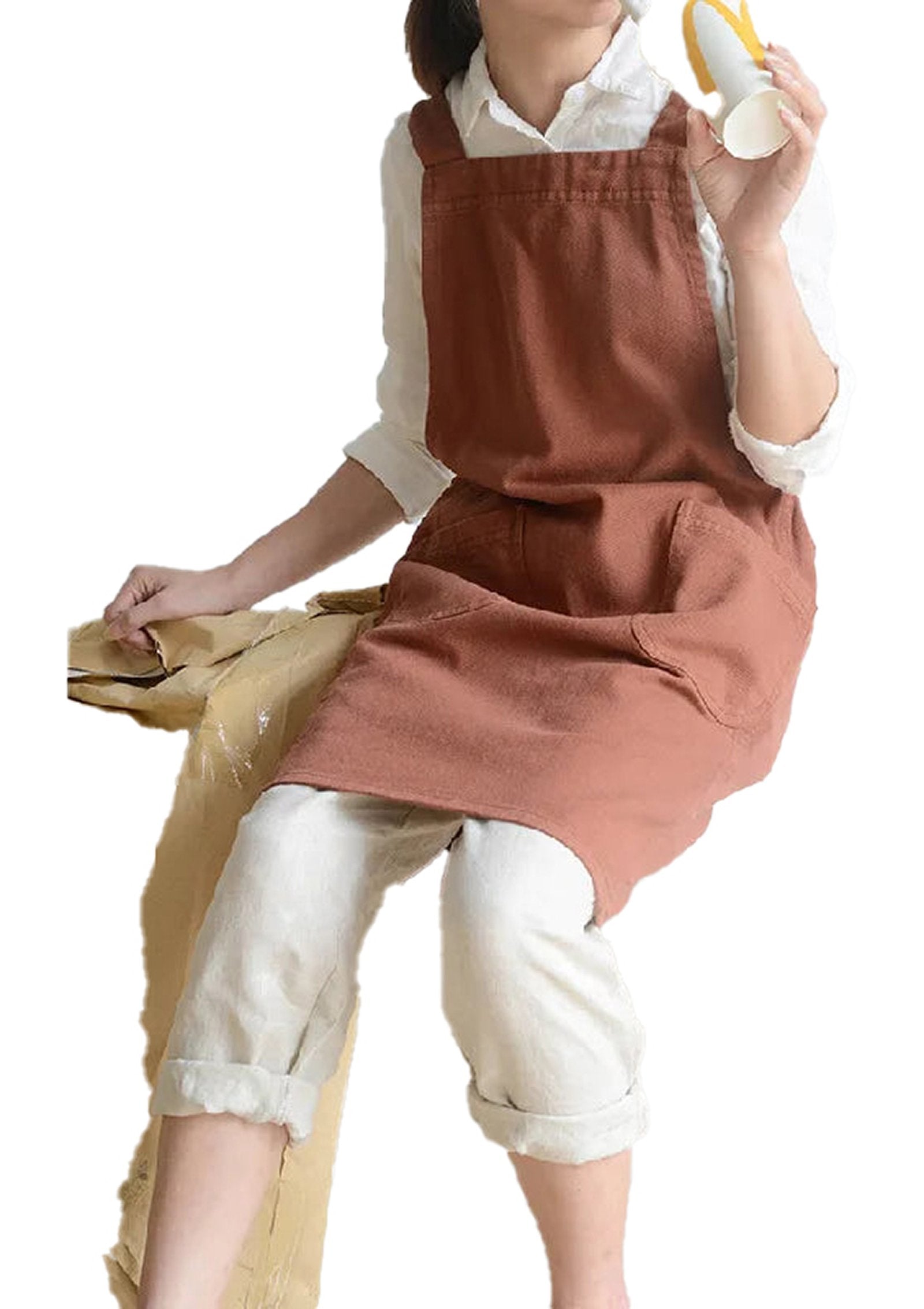 Cotton linen cross back apron in rust color worn by a model sitting, holding a cup