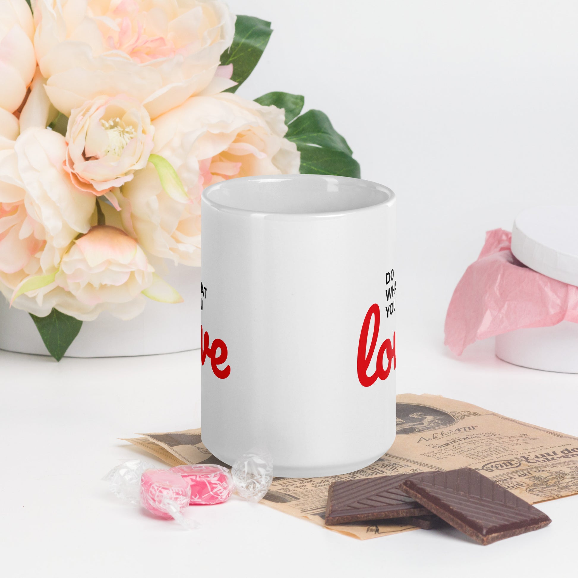 White do what you love coffee mug by LuckeLadybug with red text, placed on a newspaper with chocolates and candy nearby