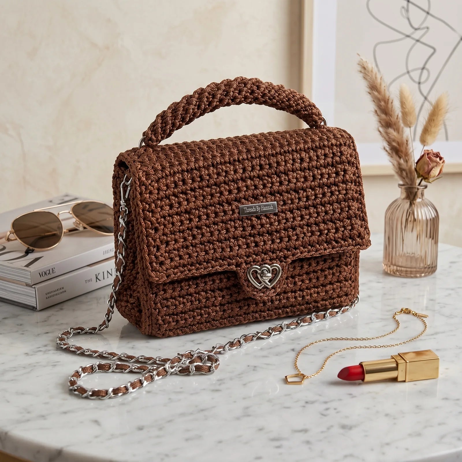 Brown crochet handbag with a chain detail on a marble surface with books and a vase in the background.