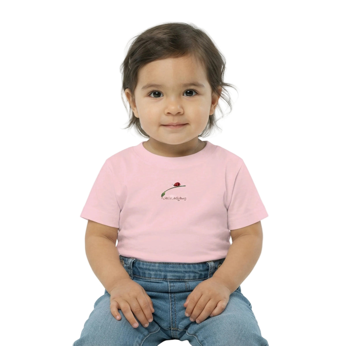 A toddler girl with dark hair sitting against a white background, wearing a light pink short-sleeve t-shirt with a small ladybug graphic and the text 'love always' on the chest, paired with blue jeans.