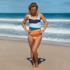 Blonde model wearing elegant striped one-piece swimsuit for women on sandy beach with sunglasses and hands on hips